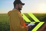 Person mit Jacke auf Feld, betrachtet Tablet. Im Hintergrund ein weiter grüner Acker bei Sonnenuntergang.
